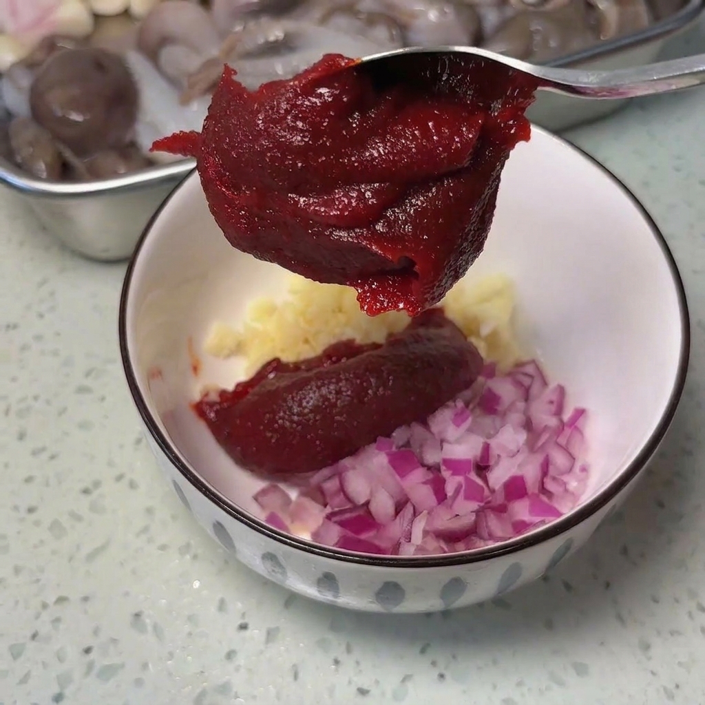 Ein Metalllöffel mit einem dicken Klecks dunkelroter koreanischer Chilipaste über einer weißen Schüssel mit Knoblauch und Zwiebeln.