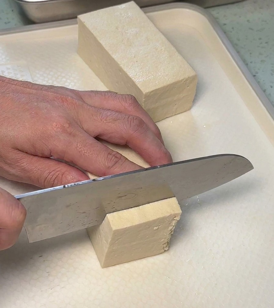 Eine Person schneidet einen Block festen weißen Tofu mit einem Kochmesser in rechteckige Stücke auf einem Schneidebrett.