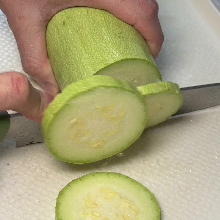 Hände schneiden eine hellgrüne Zucchini in runde Scheiben auf einem weißen Schneidebrett.