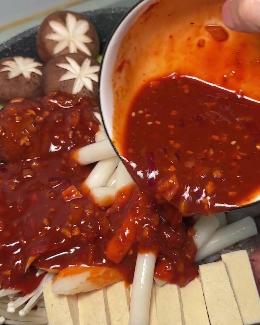 Scharfe rote Sauce wird aus einer Schüssel über die rohen Zutaten und Reiskuchen in den Kochtopf gegossen.