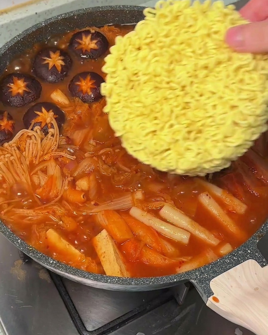 Eine Hand legt einen runden Block trockener Instant-Ramen in einen Topf mit kochender, roter Suppe.
