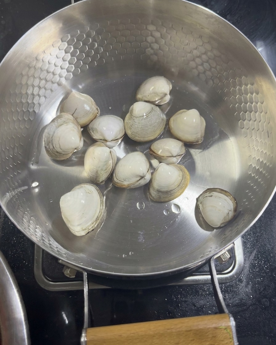 Mehrere rohe weiße Venusmuscheln liegen am Boden einer Edelstahlpfanne mit Wabenstruktur bereit zum Dämpfen.