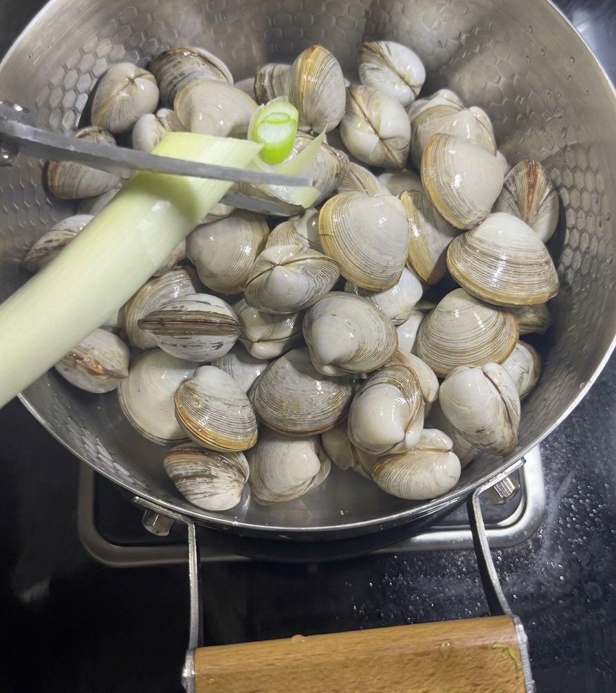 Eine Hand schneidet mit einer Schere Frühlingszwiebeln über einer Pfanne mit rohen Muscheln und Ingwerscheiben.