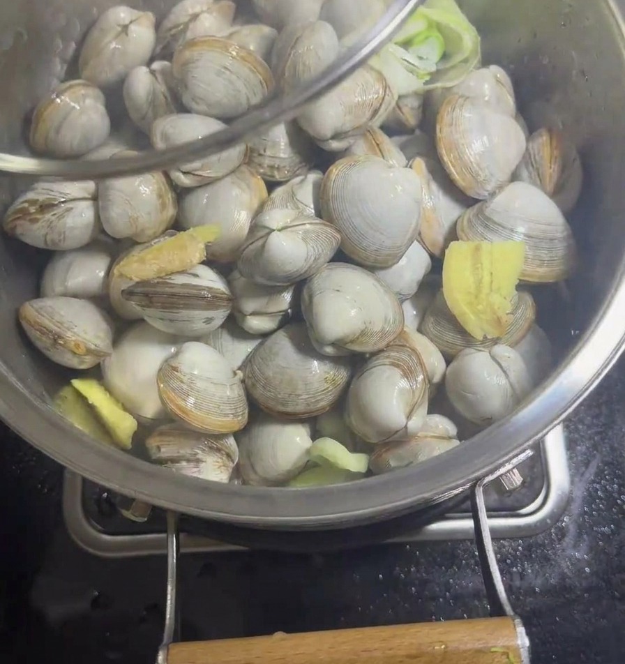 Eine Edelstahlpfanne mit Muscheln und Aromaten wird mit einem klaren Glasdeckel abgedeckt.