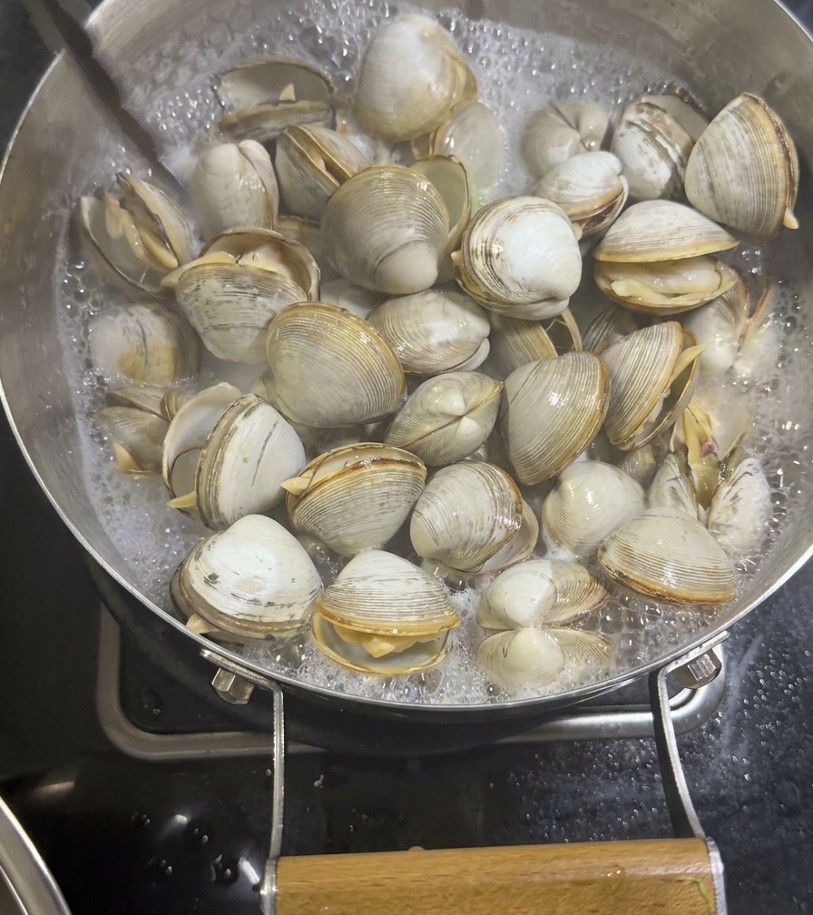 Eine Metallzange hebt geöffnete, gegarte Venusmuscheln aus einer Pfanne mit sprudelnder, milchiger Brühe.