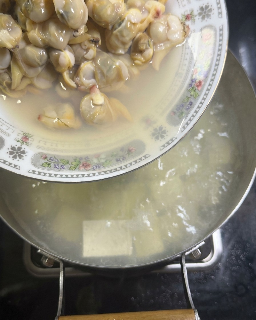 Ein Teller mit ausgelöstem Muschelfleisch wird in einen Topf mit köchelnder Brühe und Seidentofu geleert.