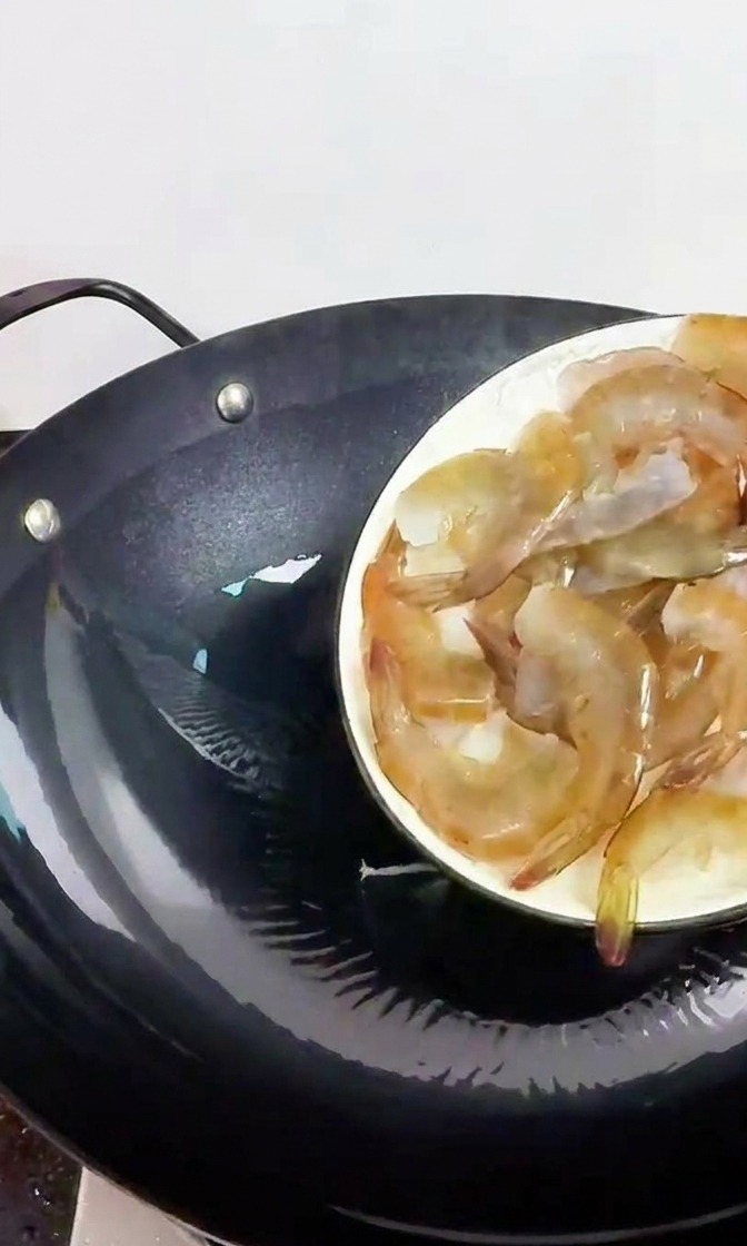 Eine Hand schiebt frische, graue Garnelen von einem weißen Teller in einen vorgeheizten schwarzen Gusseisen-Wok.