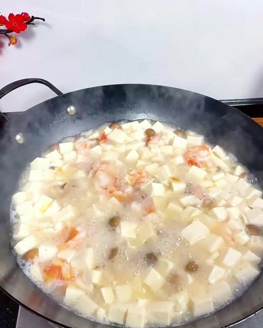 Ein Metalllöffel streut feines weißes Pfefferpulver in die sprudelnde Tofu-Meeresfrüchte-Suppe im Wok.