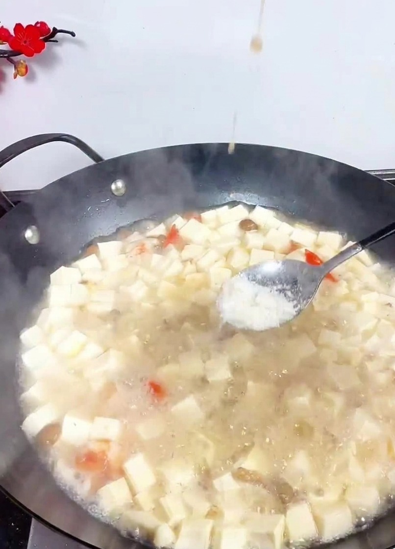 Ein Metalllöffel streut weißes Salz in einen großen Wok mit köchelndem Tofu und Meeresfrüchten in heller Brühe.