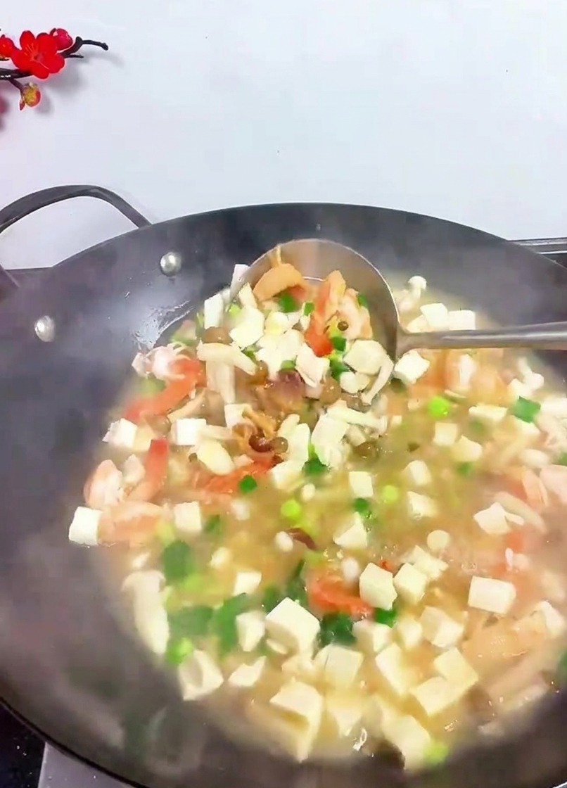 Eine große Schöpfkelle hebt und mischt Tofuwürfel, Garnelen und grüne Kräuter in einem dampfenden Wok.