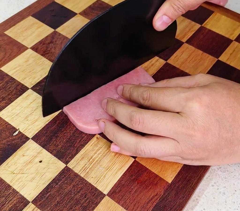 Eine Person würfelt eine pinkfarbene Schinkenwurst mit einem großen Kochmesser auf einem karierten Holzbrett.
