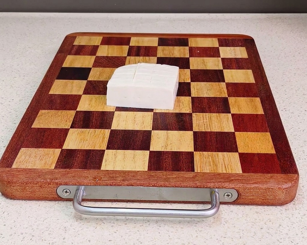 Ein Block frischer weißer Tofu, in gleichmäßige Würfel geschnitten, auf einem braun-beigen karierten Holzbrett.