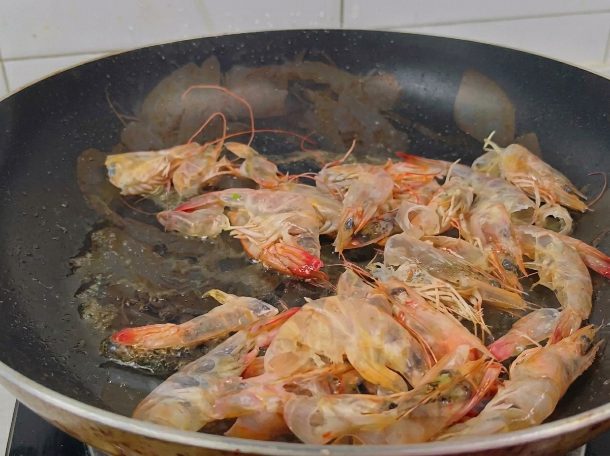 Eine schwarze Bratpfanne mit Garnelenschalen, die in glänzendem Öl angebraten werden und eine leuchtend orange Farbe annehmen.
