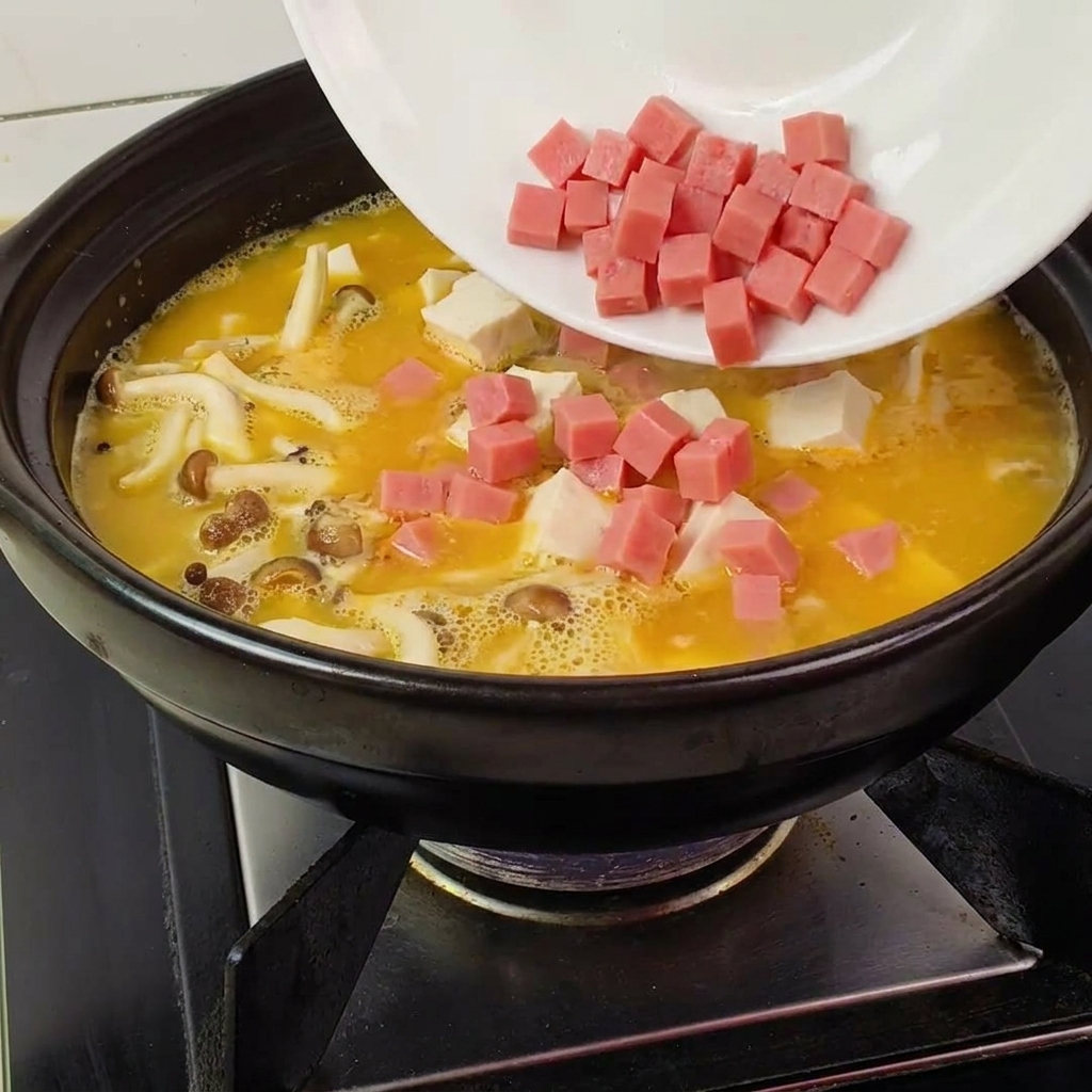 Gewürfelter Schinken wird von einem weißen Teller in den Tontopf zu gelber Brühe, Pilzen und Tofu gegeben.