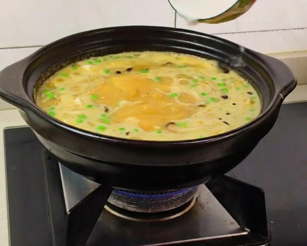 Eine Hand gießt weiße Stärkemischung aus einer Keramikschale in die sprudelnde gelbe Suppe im Tontopf.