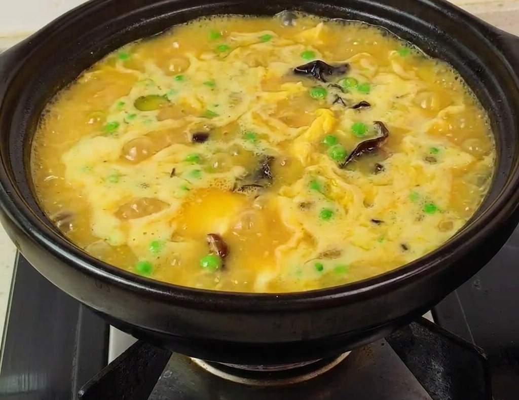 Ein Tontopf mit dicker, gelber Suppe, Garnelen, Erbsen und Pilzen auf einem brennenden Herd.