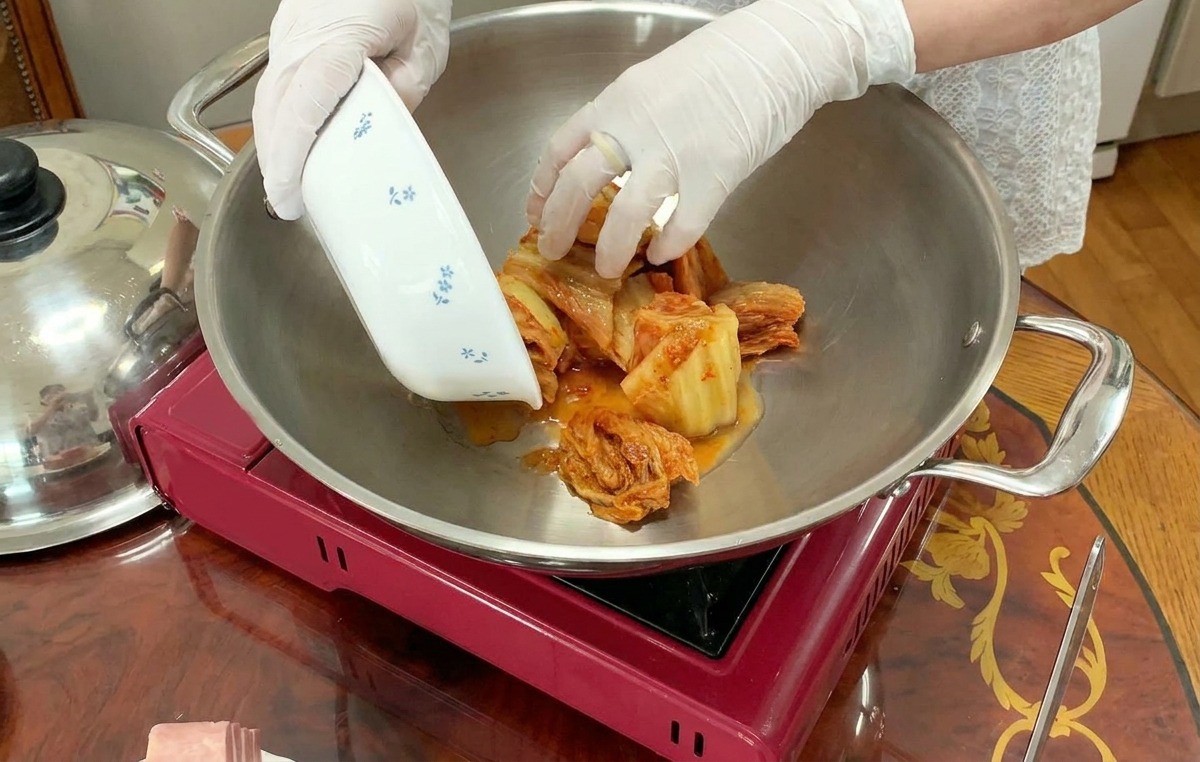 Eine Person mit weißen Handschuhen gibt geschnittenes Kimchi aus einer kleinen Schale in die Mitte eines großen Topfes auf einem Herd.