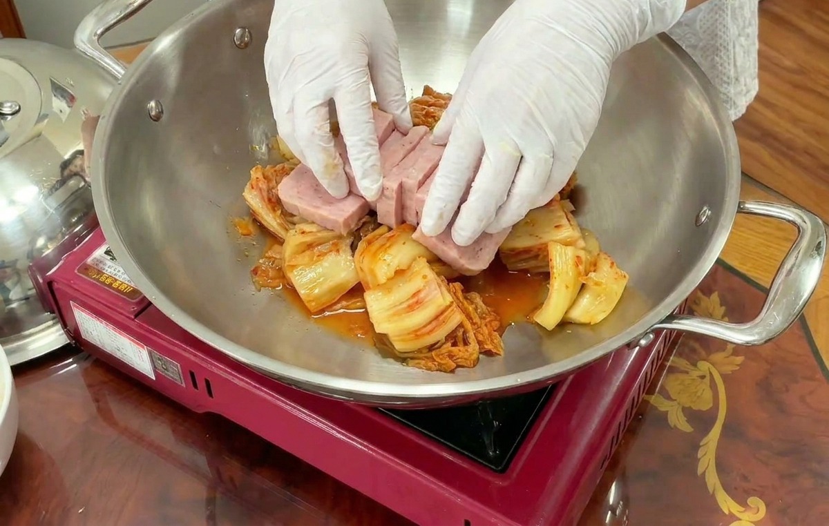Hände legen sorgfältig rechteckige Scheiben Frühstücksfleisch um einen Berg aus Kimchi in einem Topf.