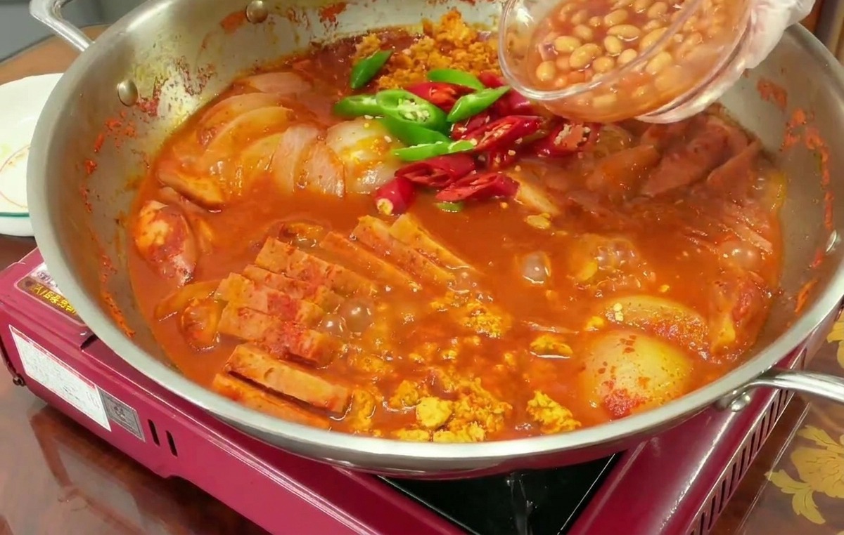 Baked Beans werden in den bereits kochenden, roten Eintopf gegeben, in dem auch Chilischoten schwimmen.