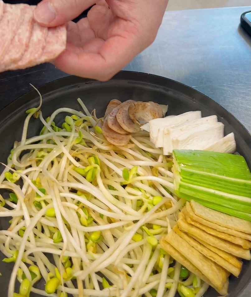 Gleichmäßige Scheiben Frühstücksfleisch werden ordentlich in den Topf neben Sprossen und andere Zutaten gelegt.