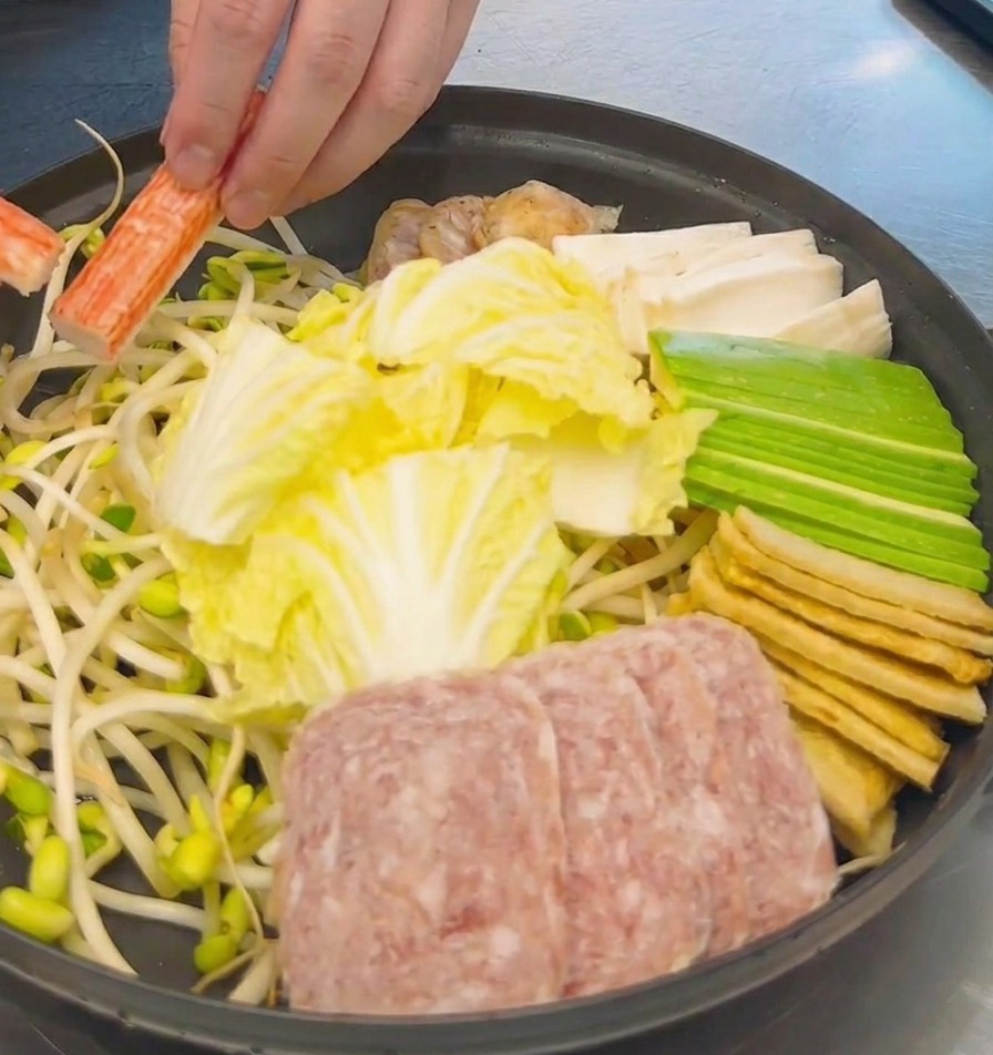 Eine Hand ordnet orange-weiße Surimi-Sticks in einem Topf zwischen Chinakohl, Fleisch und Tofu an.