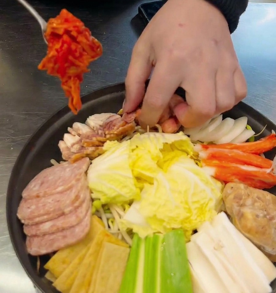 Ein Metalllöffel setzt eine Portion rotes Kimchi in die Mitte des Topfes zwischen Würstchen und Fleisch.