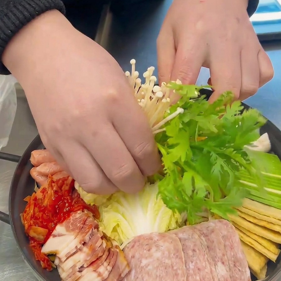 Hände platzieren Enoki-Pilze und grüne Kräuter dekorativ im Zentrum des vorbereiteten Eintopftopfs.