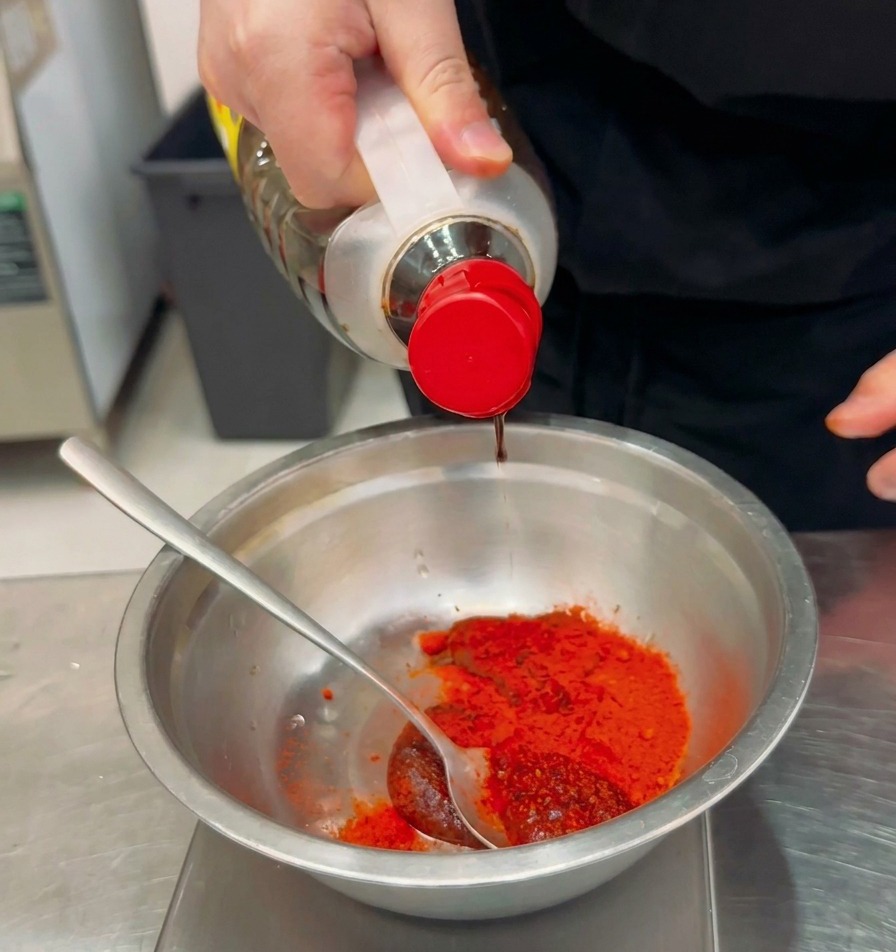Flüssige Würze wird aus einer Flasche in eine Schüssel mit rotem Chilipulver und Paste gegossen.