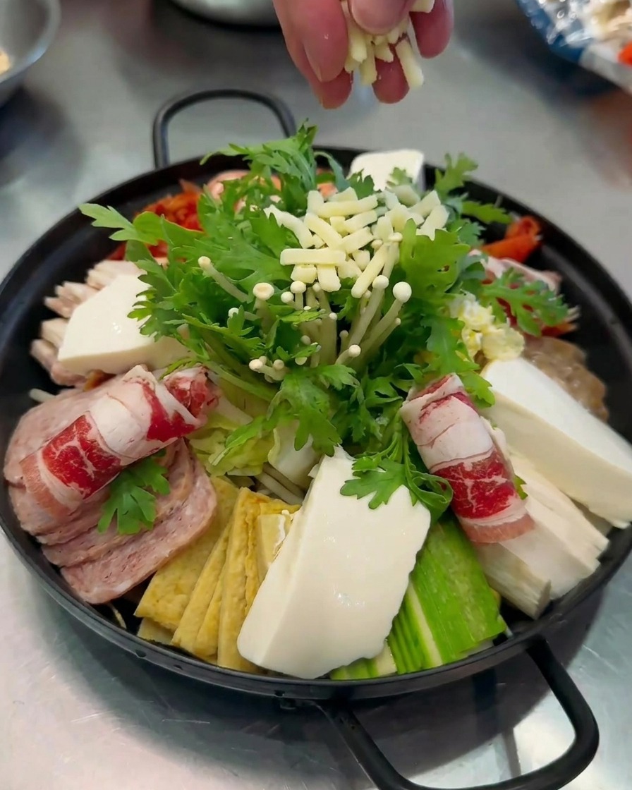 Nahaufnahme von weißem Käse, der über den fertig angerichteten und farbenfrohen Budae-jjigae-Topf gestreut wird.