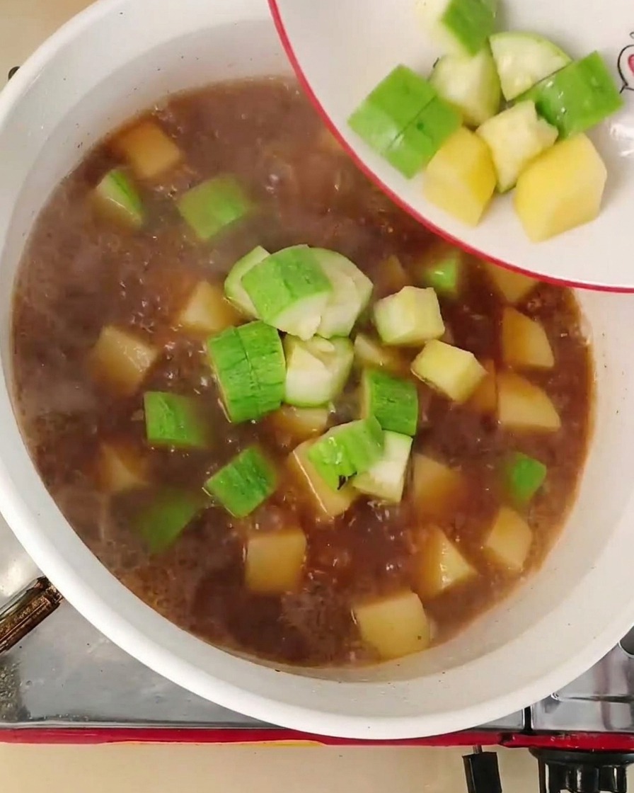 Gewürfelte Zucchini und Zwiebeln werden von einem weißen Teller in eine rotbraune Brühe geschüttet.