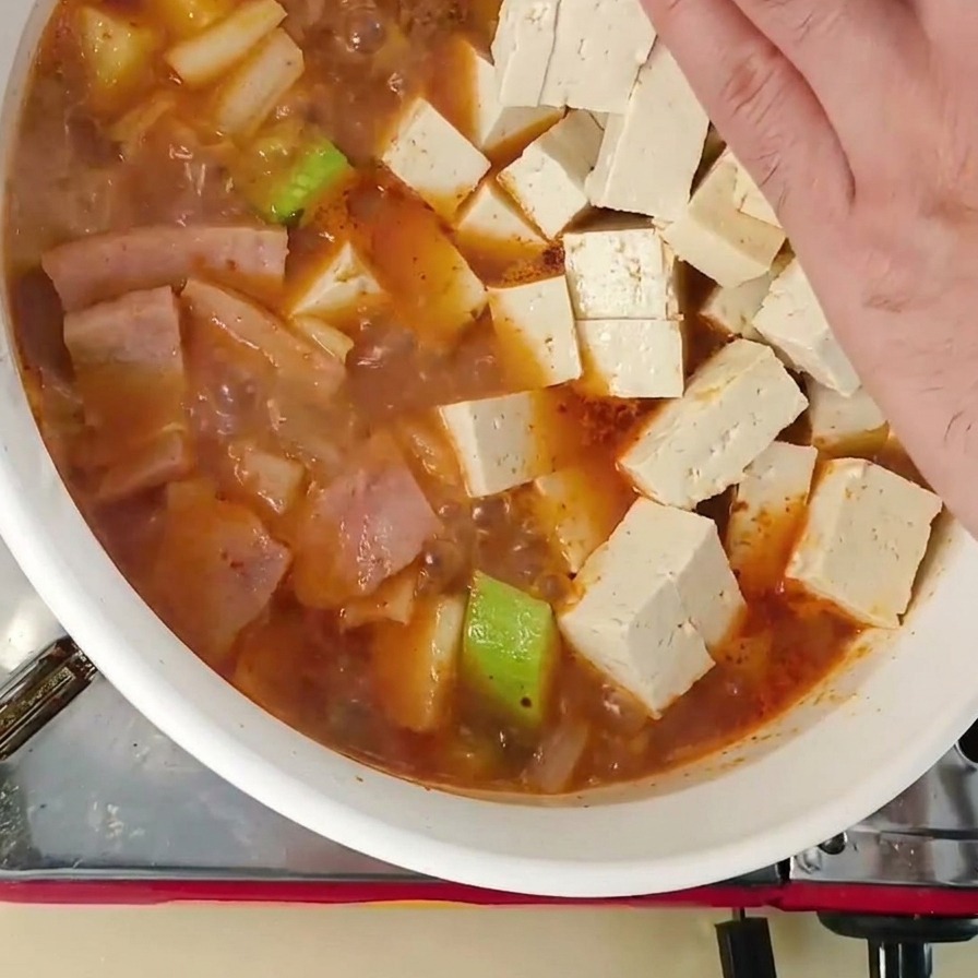 Eine Hand gibt frische weiße Tofuwürfel in den köchelnden koreanischen Eintopf.