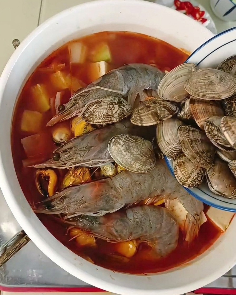 Eine kleine Schüssel schüttet frische Venusmuscheln in einen Topf mit Garnelen und Tofu.