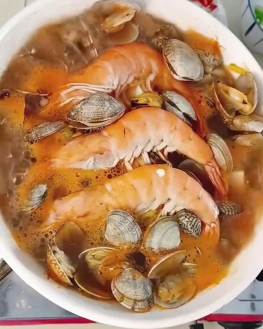 Nahaufnahme eines brodelnden Eintopfs mit Garnelen und Muscheln, garniert mit frischen Chilis und Frühlingszwiebeln.