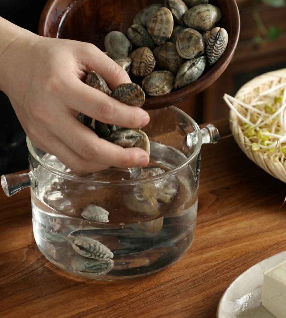 Eine Hand gießt frische Muscheln in eine Glasschale mit Wasser auf einem Holztisch.