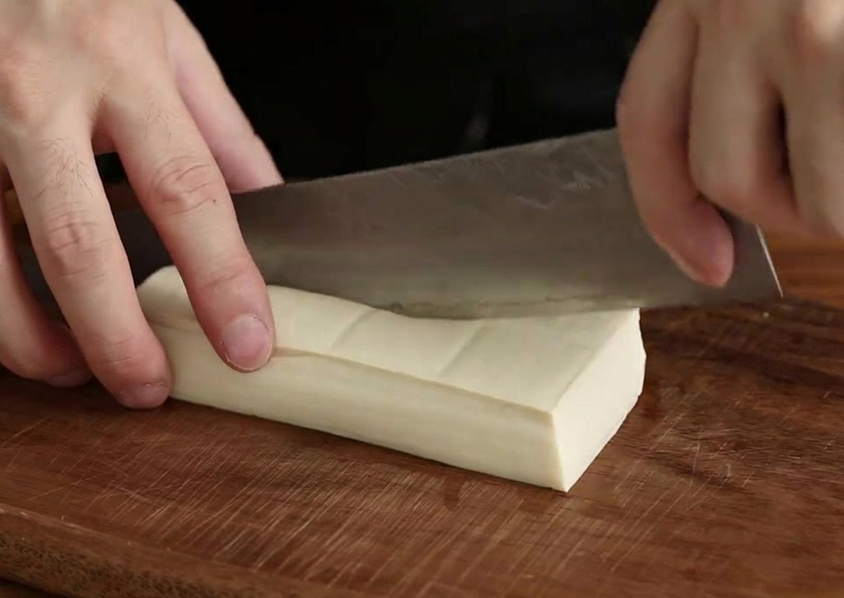 Eine Hand schneidet einen Block weißen Seidentofu mit einem Kochmesser in kleine Würfel.