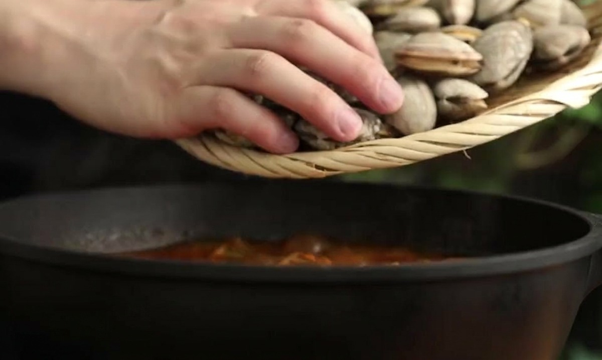 Muscheln werden mit einem kleinen Sieb in den dunklen Topf mit rotem Eintopf gegeben.