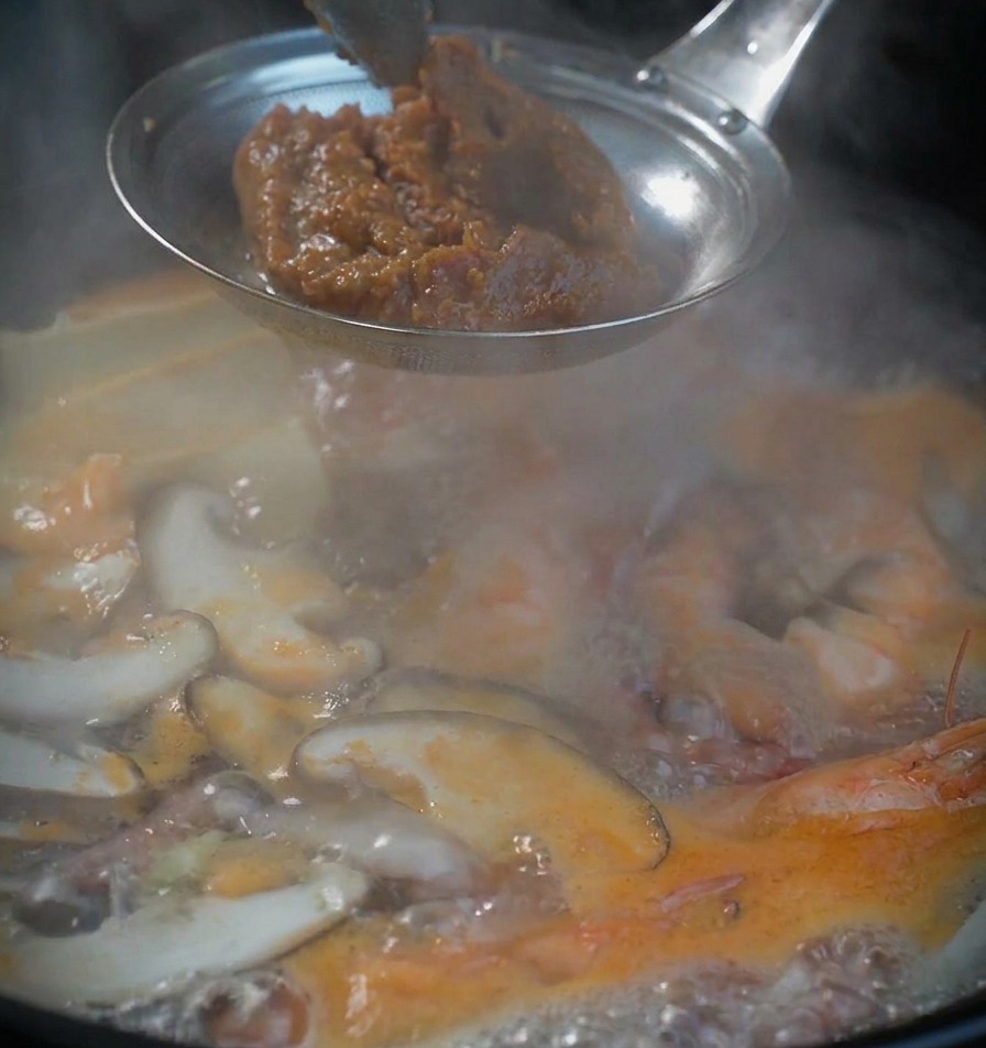 Ein Löffel dicke, strukturierte braune Sojabohnenpaste in einem Sieb über der kochenden Suppe.