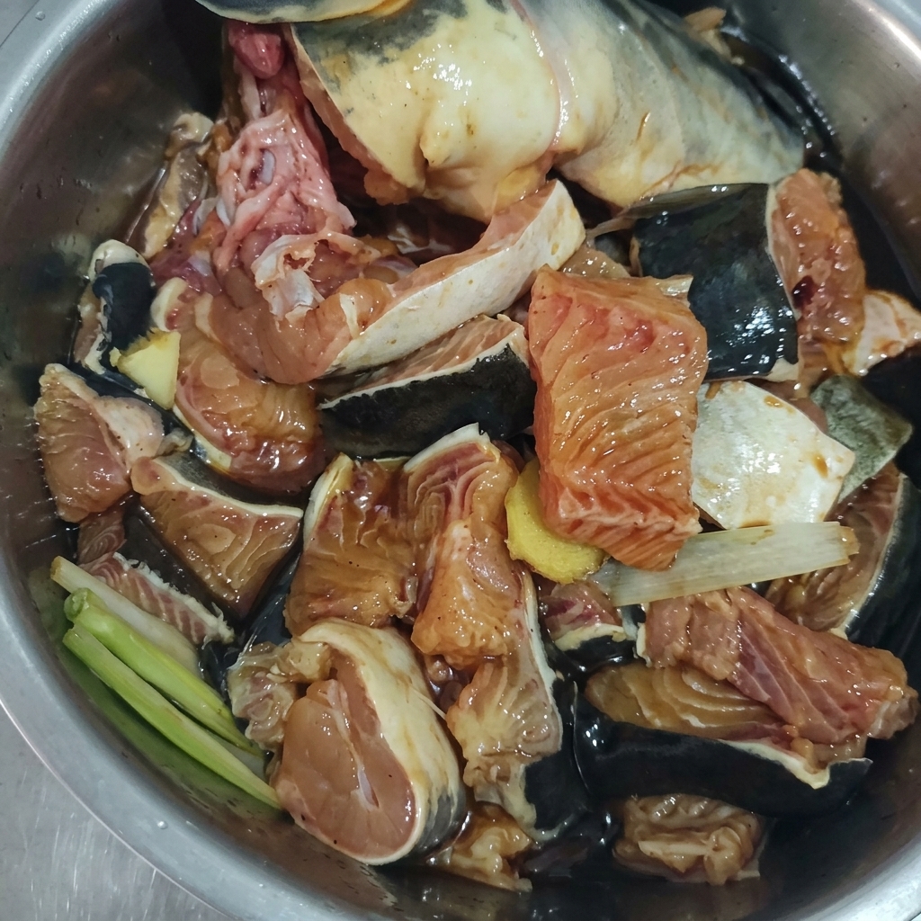 Rohe Fischstücke gemischt mit Ingwer, Frühlingszwiebeln und dunkler Marinade in einer Metallschüssel.