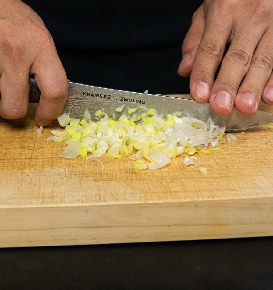 Hände schneiden Frühlingszwiebeln mit einem Kochmesser auf einem hellen Holzbrett fein.