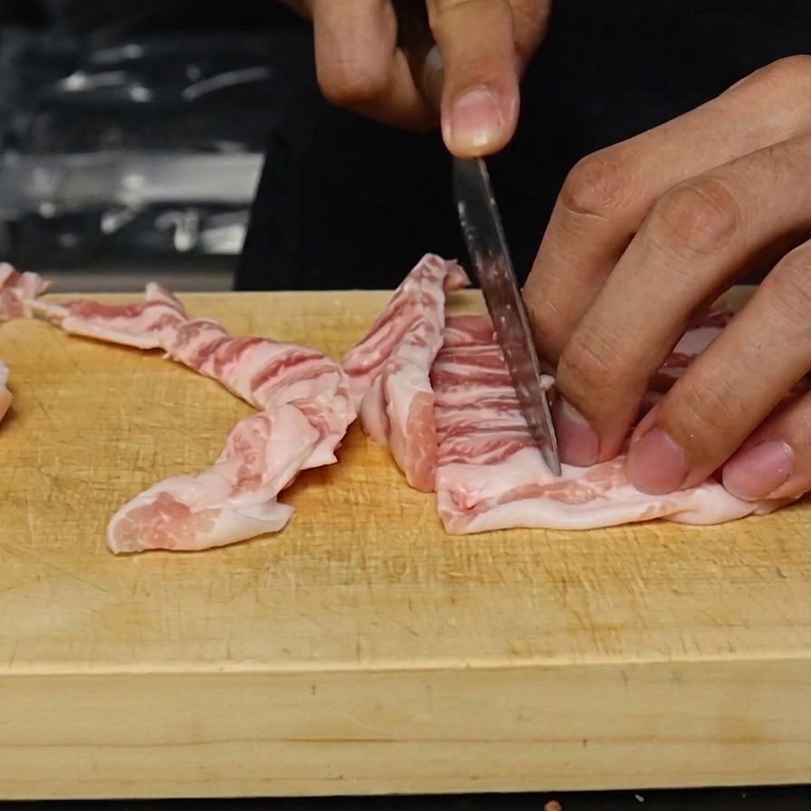 Roher Schweinebauch wird auf einem Holzbrett in etwa einen Zentimeter breite Streifen geschnitten.