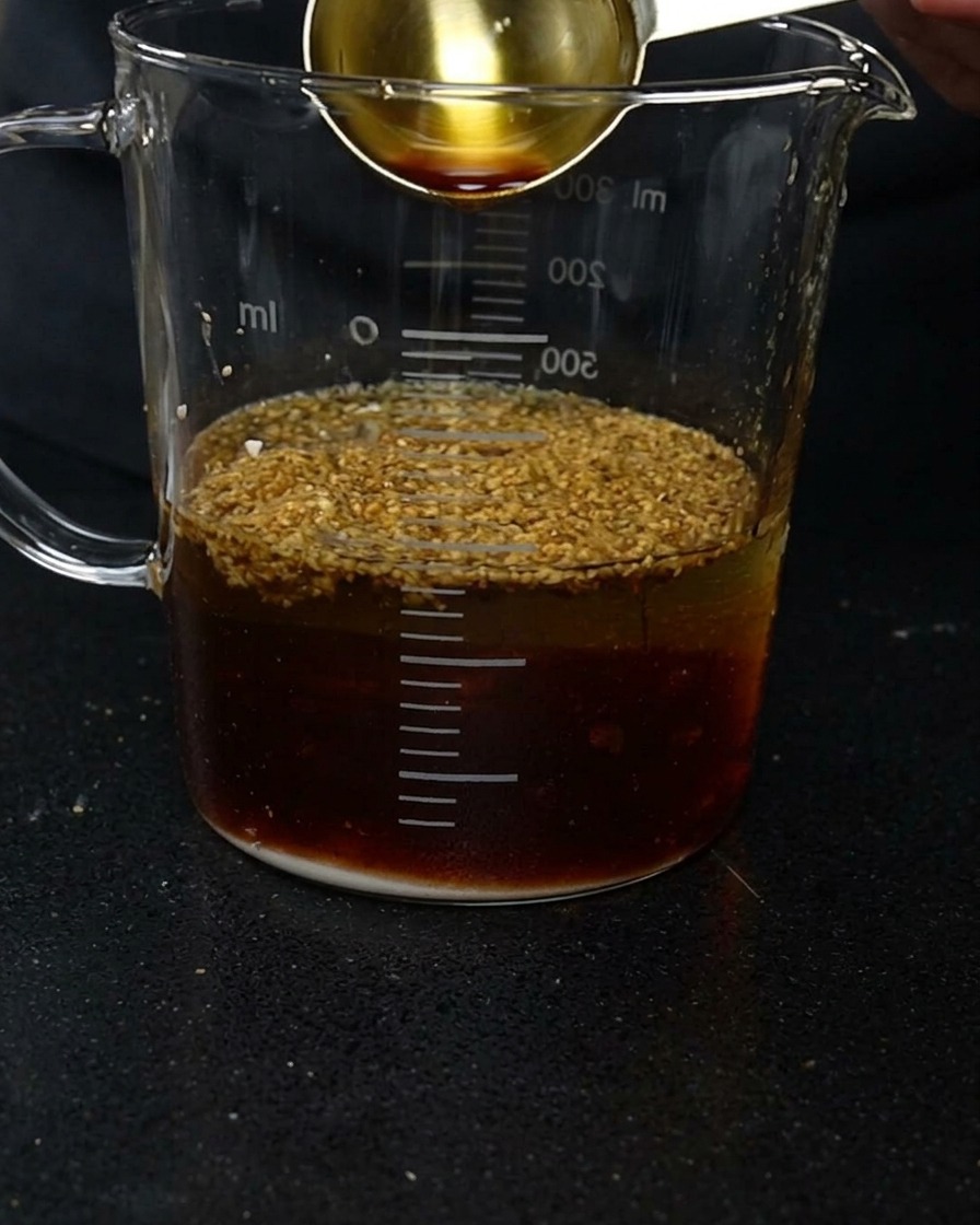 Ein goldener Messlöffel gibt dunkle Sojasauce in einen Becher mit Wasser und verschiedenen Gewürzpulvern.