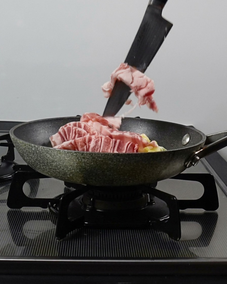 Ein Messer schiebt Schweinebauchstreifen in eine Pfanne zu den gehackten Frühlingszwiebeln und Knoblauch.