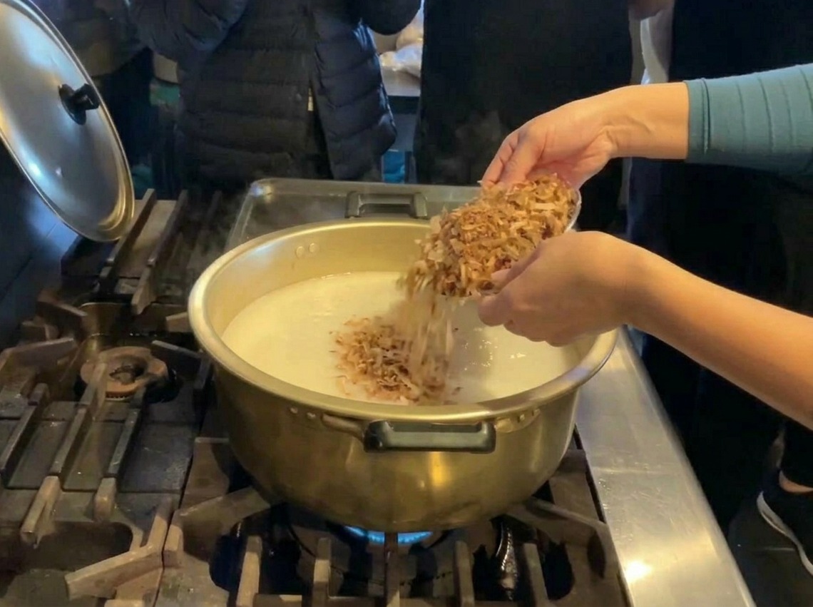 Jemand gibt eine große Menge braune getrocknete Fischflocken aus einer Silberschale in einen goldenen Topf mit weißer Brühe.