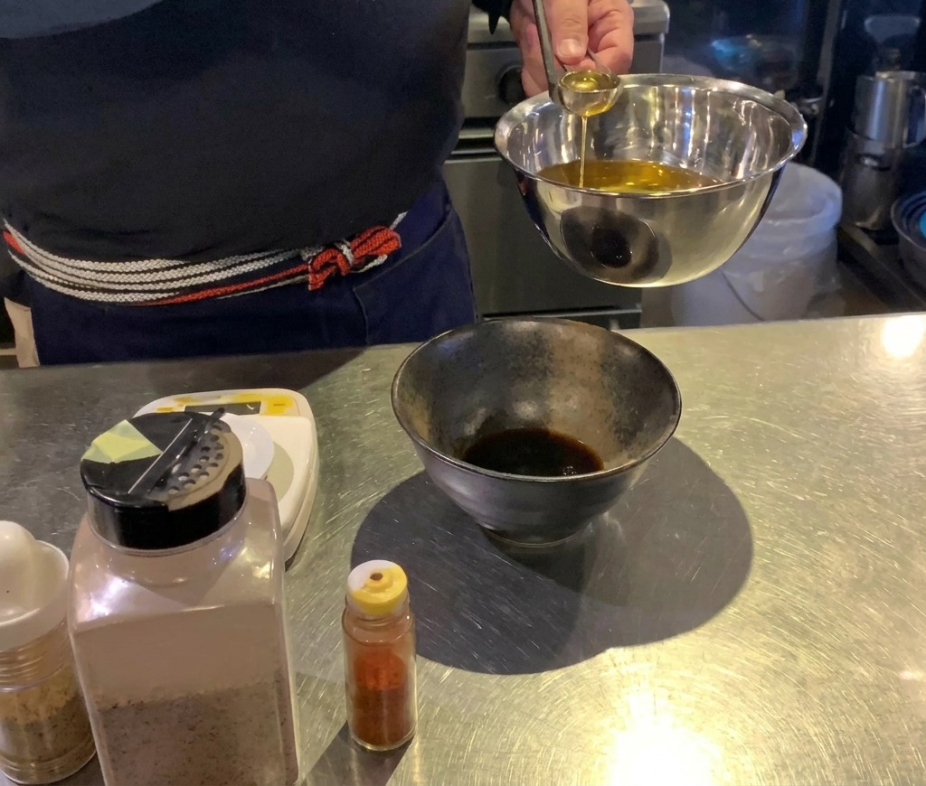 Ein Koch gibt mit einer kleinen Schöpfkelle goldenes Öl in eine schwarze Schüssel mit dunkler Sauce.