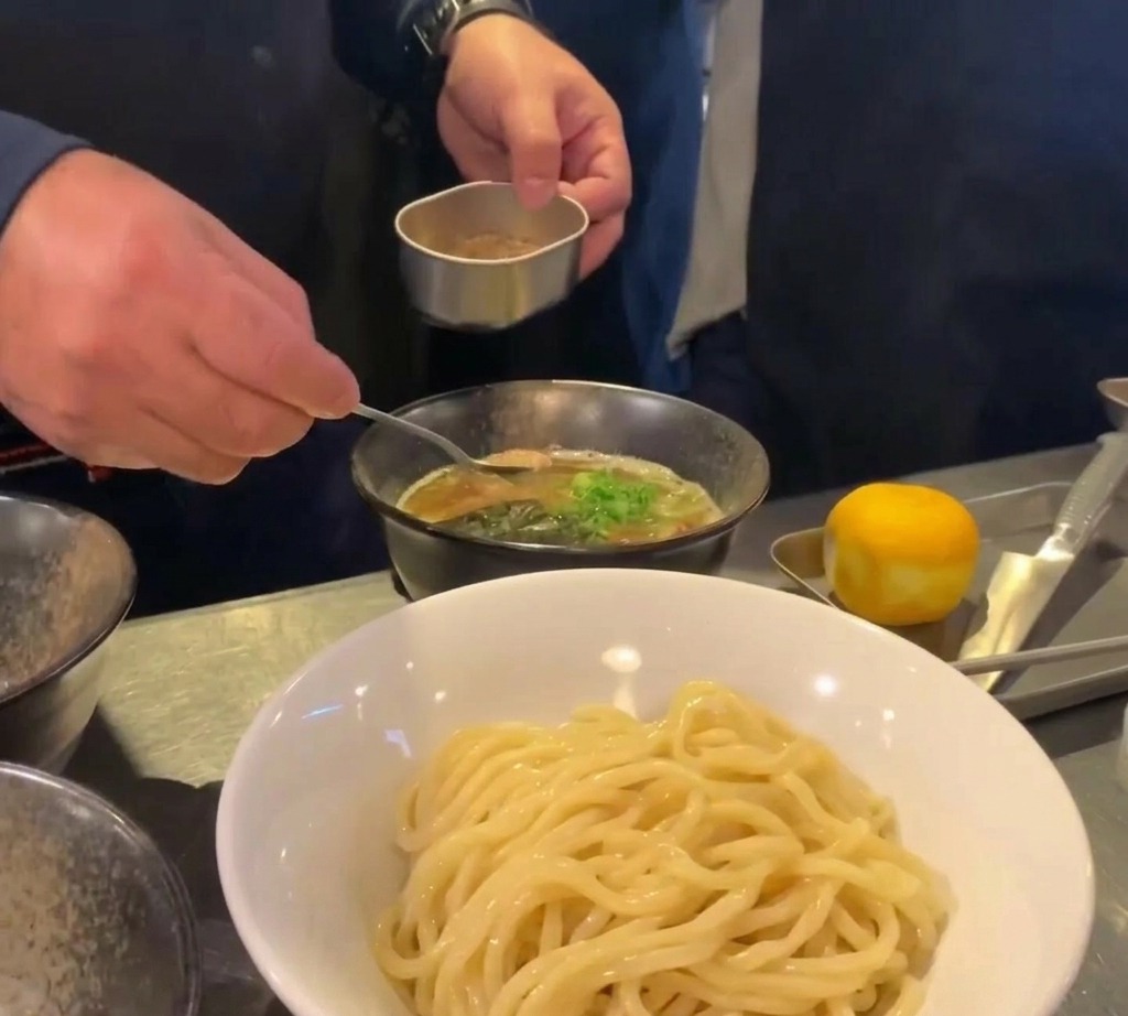 Ein kleiner Löffel platziert einen Haufen braunes Fischpulver in einer Schale mit dunkler Brühe, garniert mit Frühlingszwiebeln.