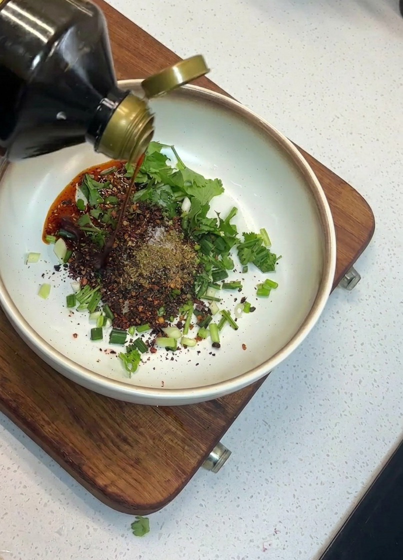 Dunkle Sojasauce wird aus einer Glasflasche in eine Schüssel mit Kräutern und Chilis gegossen.