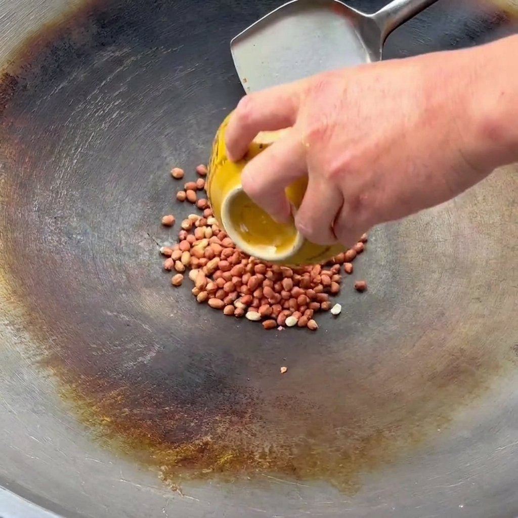 Eine Hand schüttet rohe Erdnüsse aus einer gelben Schüssel in einen Wok, in dem ein Metallspatel zum Rühren bereitliegt.