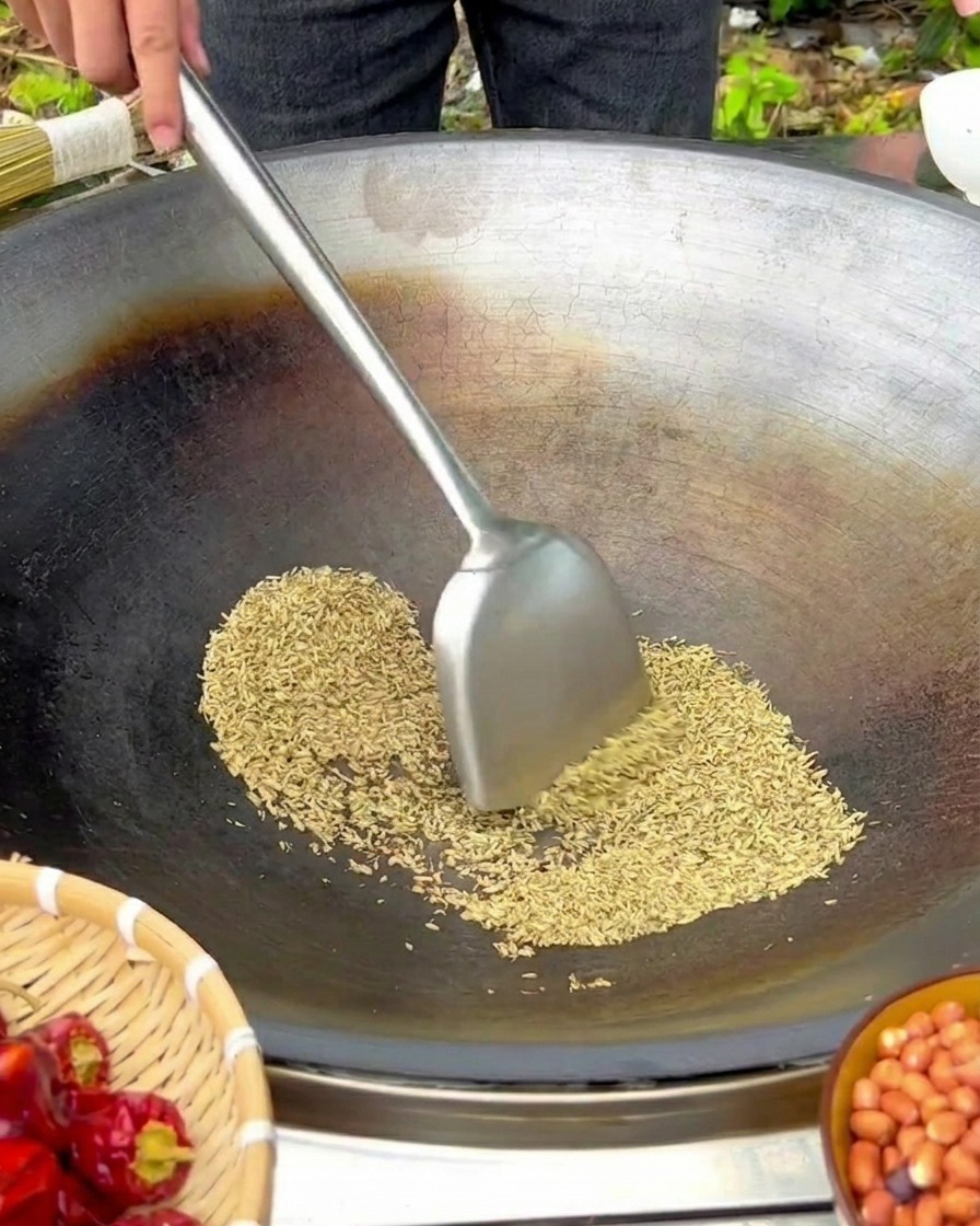 Fenchelsamen werden mit einem Metallspatel in einem großen, gusseisernen Wok über niedriger Flamme gerührt.