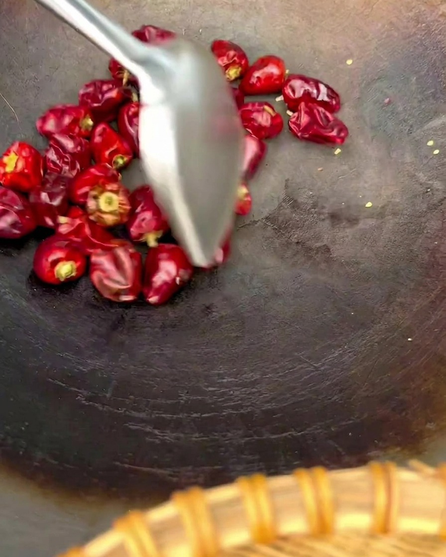 Ein Metallspatel rührt eine Handvoll runder roter Laternen-Chilis in einem dunklen Wok um.