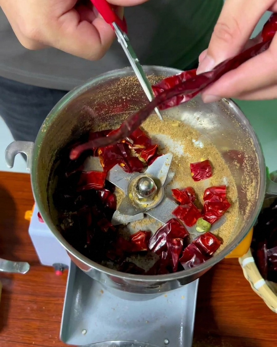 Hände benutzen eine Schere mit roten Griffen, um lange getrocknete rote Chilis in den Behälter einer Edelstahl-Mühle zu schneiden.